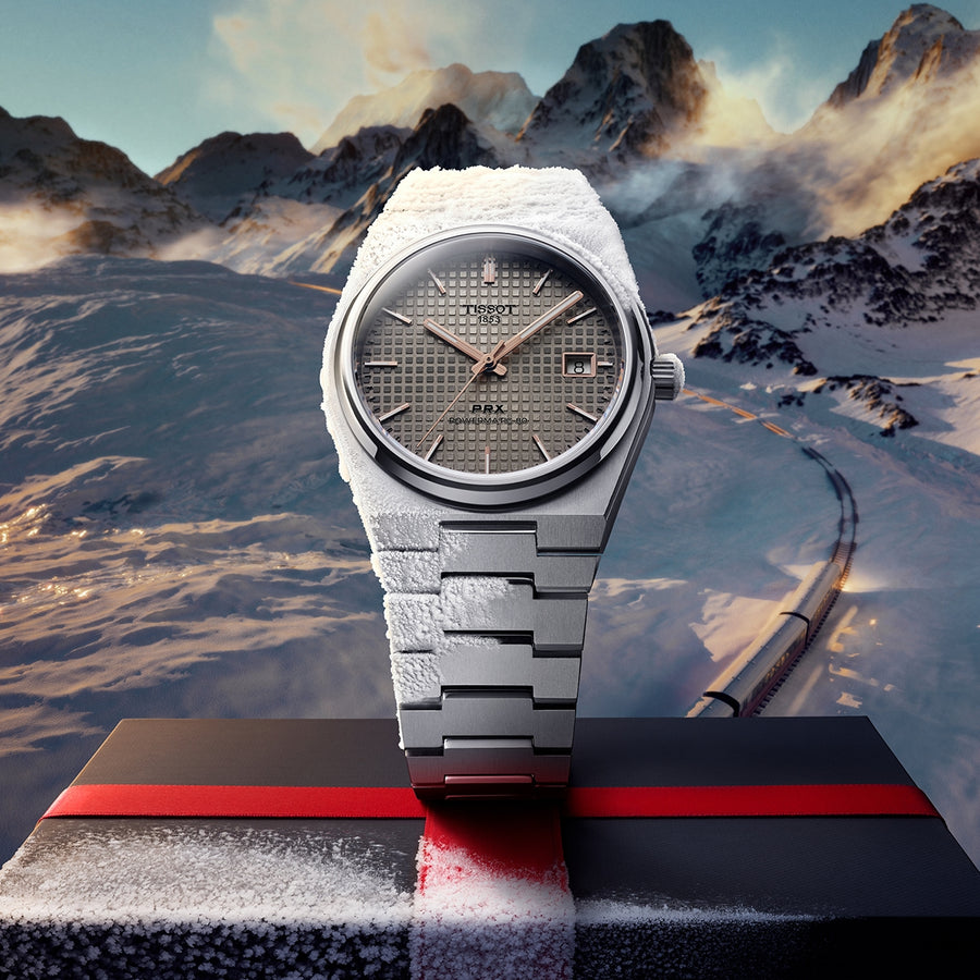 Tissot PRX givrée, cadran gris gaufré, sur une boîte noire à bande rouge, montagnes enneigées et voie ferrée derrière
